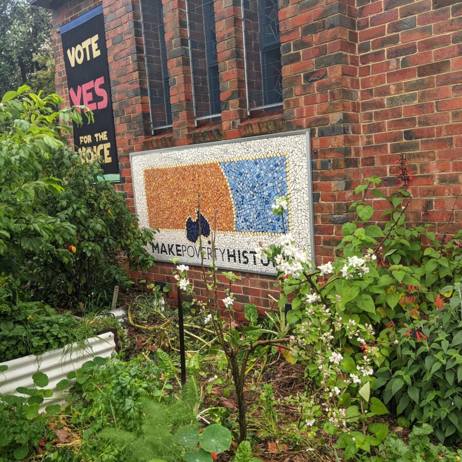 An orange and blue mozaic for "Make Poverty History" is fixed to the red-bricked, exterior wall of Fairfield Uniting Church. A "Vote Yes for the Voice" banner is on the back wall in the background.